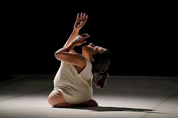 Tänzerin kniet in weißem Outfit im Scheinwerferlicht, Arme über dem Kopf erhoben, Tanzperformance von Chiara Bersani "Seeking Unicorns"